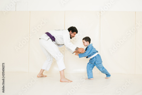 Father and little kid son are engaged in wrestling jiu-jitsu in the gym in a kimono. Trainer teaches child the methods and positions of single combat, karate or aikido. © uv_group