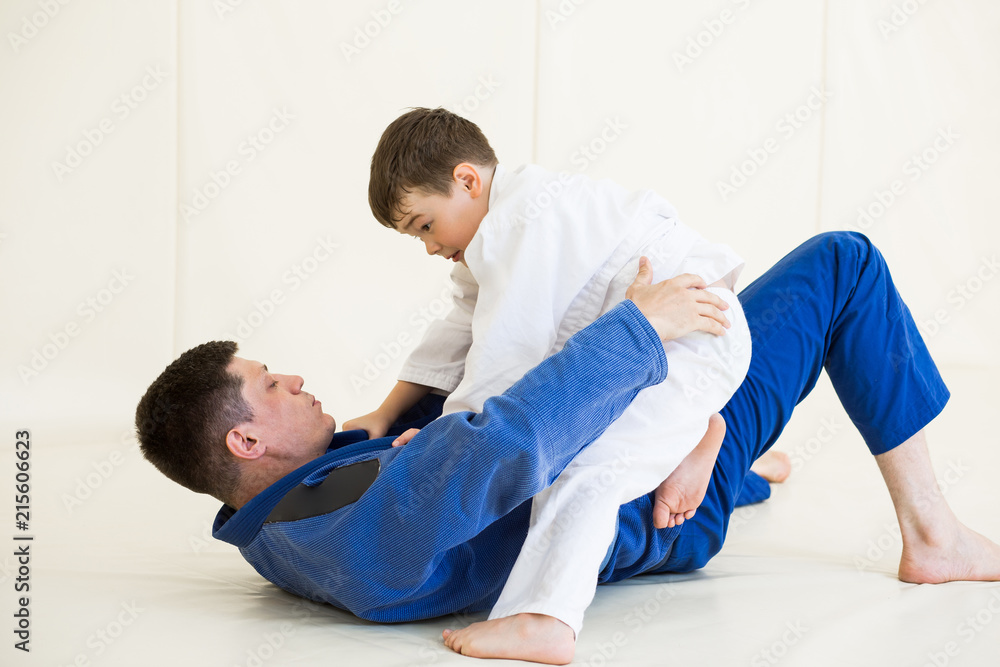 Father and little kid son are engaged in wrestling jiu-jitsu in the gym in  a kimono. Trainer teaches child the methods and positions of single combat,  karate or aikido. Stock Photo