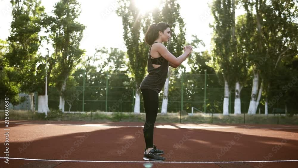 Young woman athlete in sports clothes squat exercise outdoor. Spectacular brunette in black T-shirt and legging enthusiastically doing beloved physical exercise. Side view Young woman athlete in sports clothes squat exercise outdoor. Spectacular brunette in black T-shirt and legging enthusiastically doing beloved physical exercise. Side view