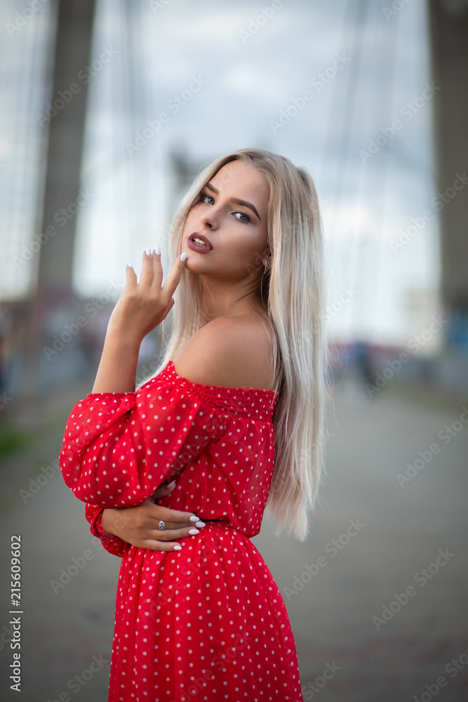 Adorable blonde model wearing fashionable red dress, posing on the bridge in the evening