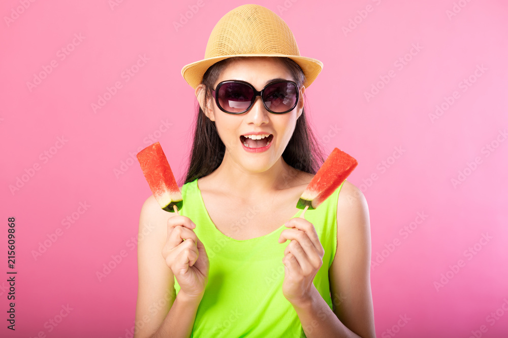 Portrait of a happy attractive woman in summer green outfit with hat and sunglasses holding fresh water melon on stick isolated over pink background. Summer vibe concept. Portrait of a happy attractive woman in summer green outfit with hat and sunglasses holding fresh water melon on stick isolated over pink background. Summer vibe concept.