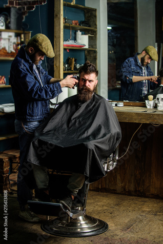 Hipster client getting haircut. Barber with clipper trimming hair on temple of client. Barber with hair clipper works on hairstyle for bearded man barbershop background. Hipster lifestyle concept