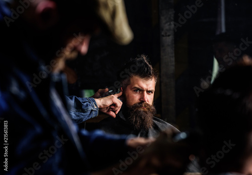Barber with hair clipper works on hairstyle for man with beard, barbershop background. Barber styling hair of brutal bearded client with clipper. Haircut concept. Hipster client getting haircut