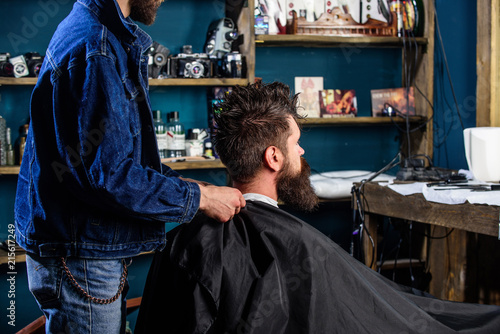 Man with beard and mustache sits in barbershop, beauty supplies on background. Barbershop concept. Man bearded client of hipster barbershop. Hipster with beard waits while barber cover neck with cape