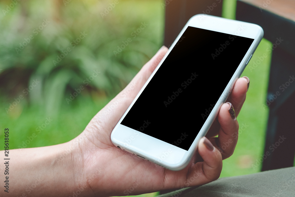 Mockup image of a woman holding white smart phone with blank black desktop screen with blur green nature background