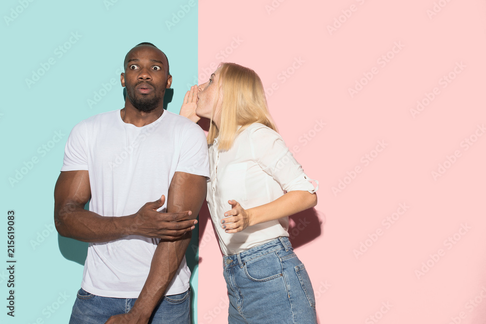 The young couple whispering a secret behind her hand over studio background The young couple whispering a secret behind her hand over studio background