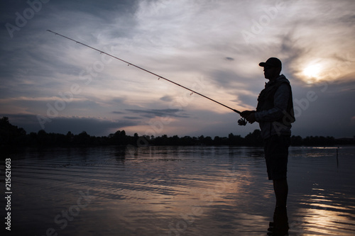 It is evening outside. Fisherman stands in shallow and fishing. He holds fly rod in hands. There is a reel under it. He stands without moving.