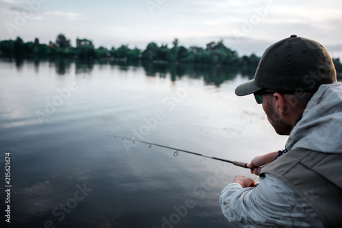 Cut view of man standing with his back to camera. He is calm and concentrated. Guy is looking at the water. He holds fly rod with both hands. It is evening outside.