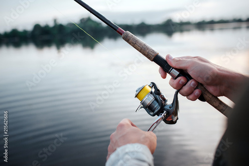 A picture of rotative spinner that guy holds in hands. He rotates reel with left hand. He is at lake. It is evening outside.