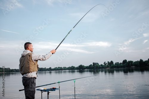 Fisherman is standing near watr and holding fly rod with right hand. He has it up really high. Man looks straight forward. Another fish rod is lying in hook.