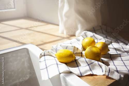 Close up shot of yellow lemons drying on checkered towel in rustic kitchen with sun shining through window. Picture of ripe citrus fruits lying on wooden counter by white sink. Selective focus