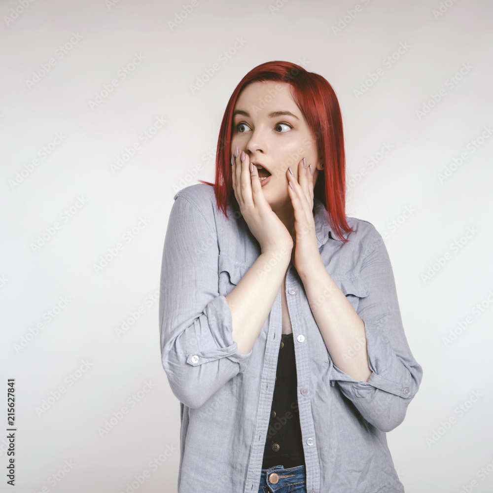 Red haired plus size ordinary woman standing on a neutral grey background. Emotional portrait. She looks scared Red haired plus size ordinary woman standing on a neutral grey background. Emotional portrait. She looks scared