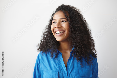 Positive human facial expressions, emotions, reaction and feelings. Attractive young dark skinned female with voluminous hairstyle laughing at joke, looking away, showing white perfect teeth
