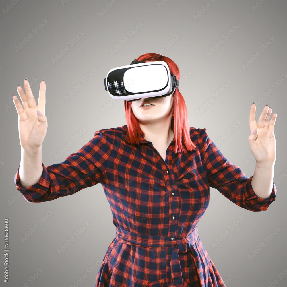 Red haired plus size ordinary woman standing on a neutral grey background. Emotional portrait. She is in a virtual reality, using VR glasses Red haired plus size ordinary woman standing on a neutral grey background. Emotional portrait. She is in a virtual reality, using VR glasses