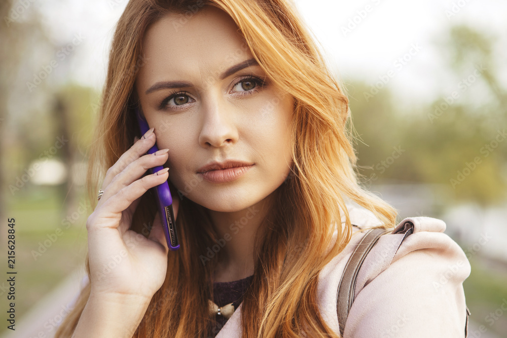 Red haired sexy plus size woman walking on a park. Cold weather, she wears leather jacket. She works on a phone, making calls