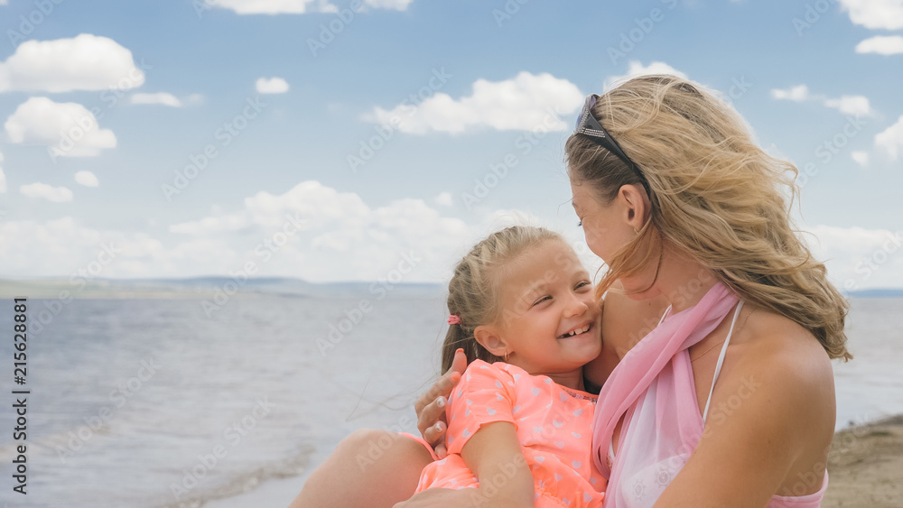 Little cute girl baby and young mother at beach. Mom sits on the shore, and her daughter is coming from behind. The kid hugs and kisses her mother. Mother and her child. Little cute girl baby and young mother at beach. Mom sits on the shore, and her daughter is coming from behind. The kid hugs and kisses her mother. Mother and her child.