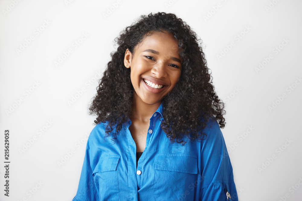 Naklejka premium Picture of gorgeous African student girl wearing casual blue shirt rejoicing at good results on exams, smiling broadly. Beautiful young black woman looking at camera with charming cute smile