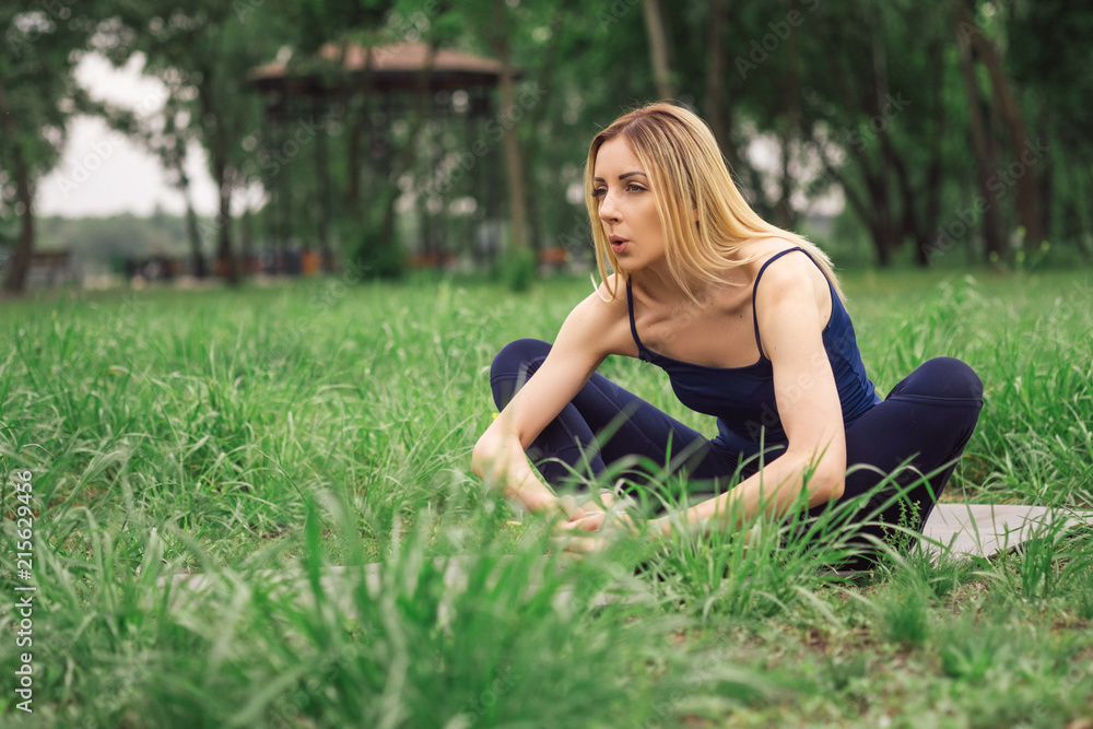 Blonde caucasian woman make stretching exercise in city park. She wears bue sport outfit.
