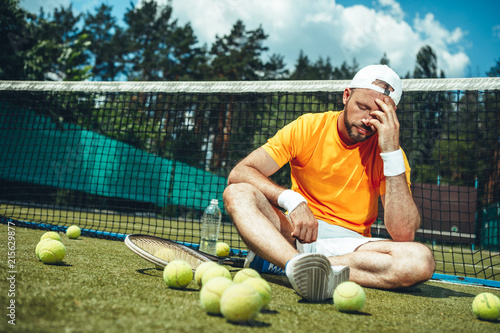 Full length portrait of bearded sad tired male athlete situating on court near net, balls and racket. He feeling headache. Exhausted sportsman after training concept photo