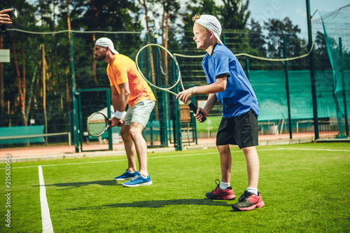 Full length side view happy man and beaming child playing tennis while situating on modern field with green grass © Yakobchuk Olena