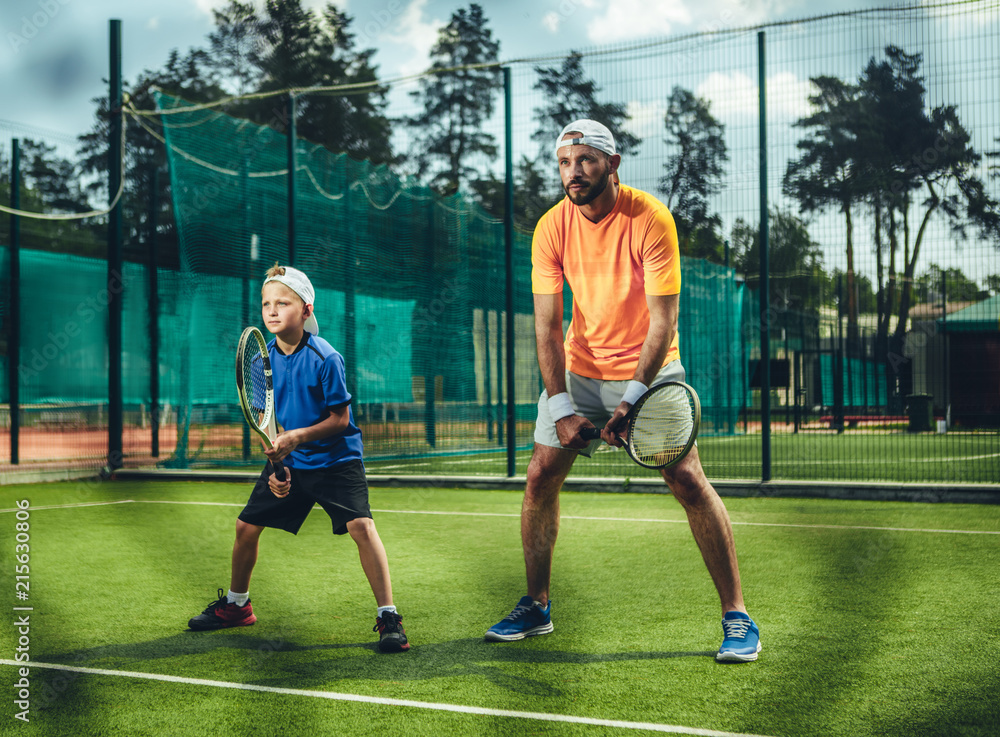 Obraz premium Full length portrait of concentrated bearded male couch playing with focused kid on contemporary court. They keeping rackets in hands