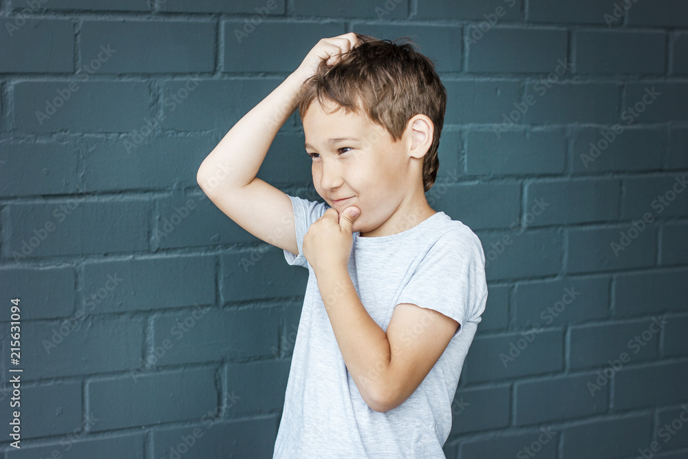 Boy 8 years old with freckles looking into the distance, thinking and scratching his head against the gray brick wall Boy 8 years old with freckles looking into the distance, thinking and scratching his head against the gray brick wall