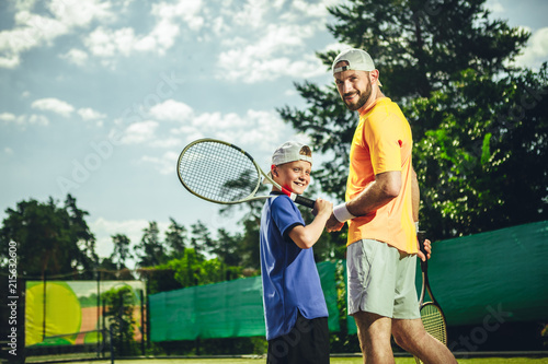 Portrait of beaming bearded man trainer wearing modern cap and cheerful child keeping sport equipment in arms. They situating outdoor © Yakobchuk Olena
