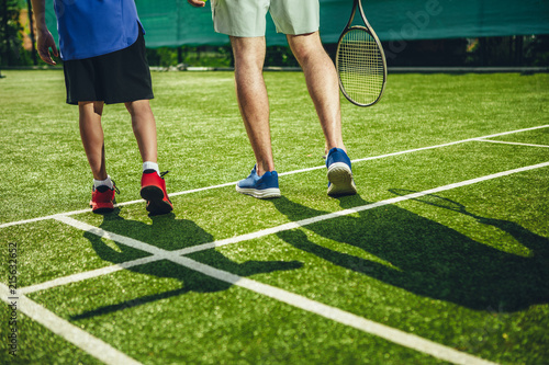 Close up male and boy legs going on field while holding rackets in arms. They playing tennis while turning back to camera