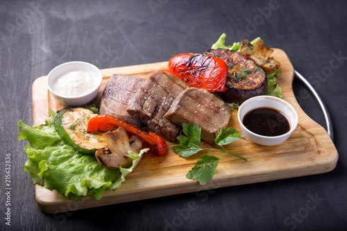 Grilled beef tongue served with vegetables and sauce on wooden board photo