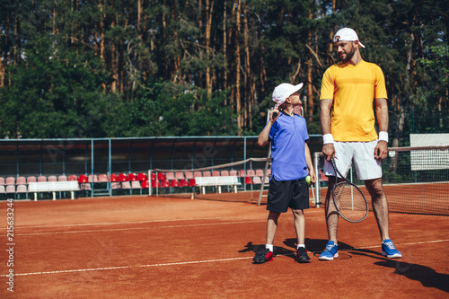 Full length side view glad little boy and beaming bearded man having fun on court outside. Satisfied guy holding ball