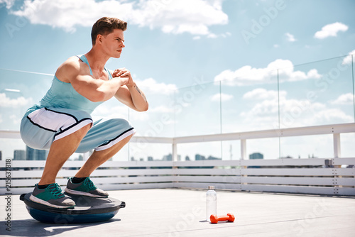 Athlete is doing sit-ups and balancing on BOSU ball. He is exercising on sunny terrace in skies above city. Copy space in right side photo