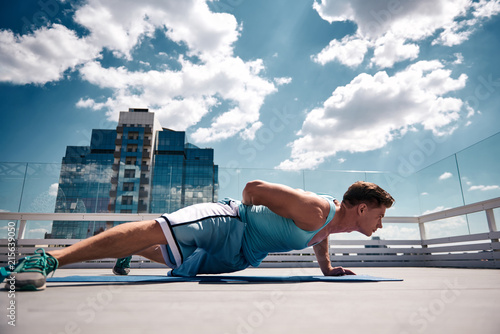 Shapely male is exercising on mat with his own weight on sunny roof in city center. He is doing push-ups while balancing on one hand. Guy is straining all body while making effort in open air