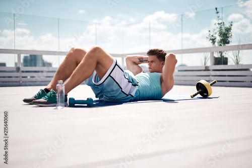 Fit guy is doing abs crunches on mat in terrace of urban building. He is spending time exercising in open air with sport equipment and water. Work out outdoors on sunny day concept