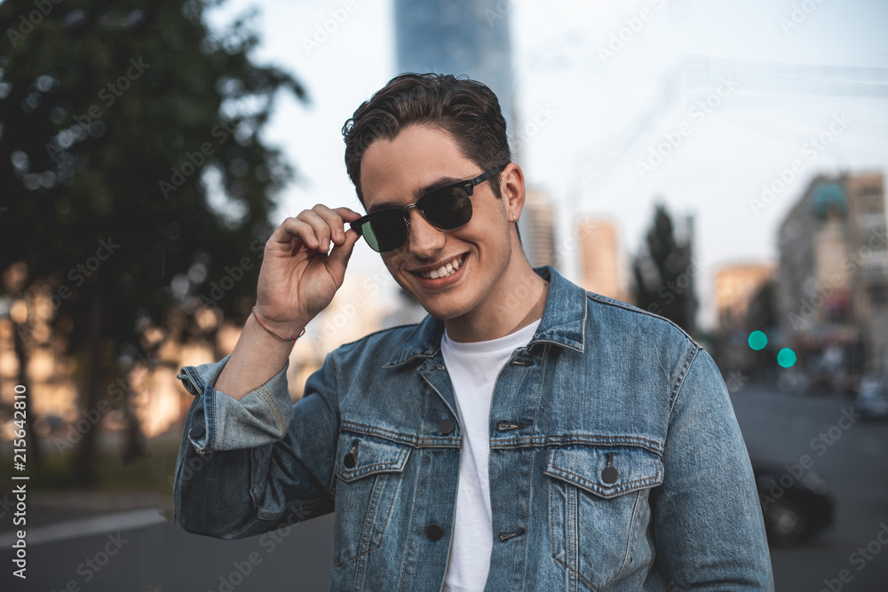 Waist up portrait of smiling young attractive man standing on street. He is touching rim of sunglasses with delight Waist up portrait of smiling young attractive man standing on street. He is touching rim of sunglasses with delight