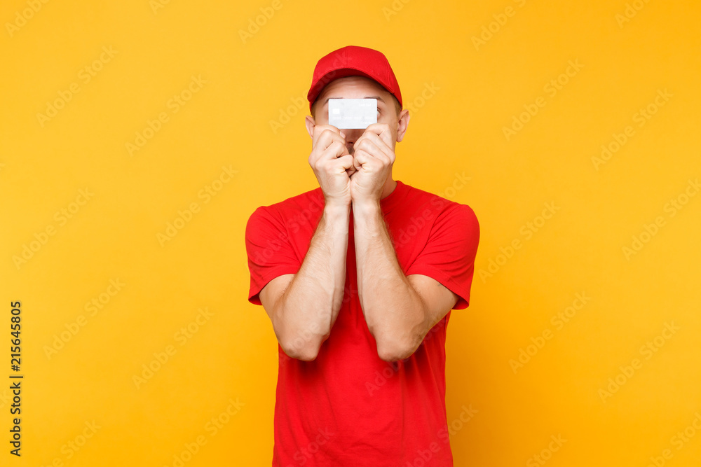 Delivery man in red uniform isolated on yellow orange background. Professional male employee in cap, empty t-shirt courier dealer hold in hand cover face credit bank card. Service concept. Copy space. Delivery man in red uniform isolated on yellow orange background. Professional male employee in cap, empty t-shirt courier dealer hold in hand cover face credit bank card. Service concept. Copy space.