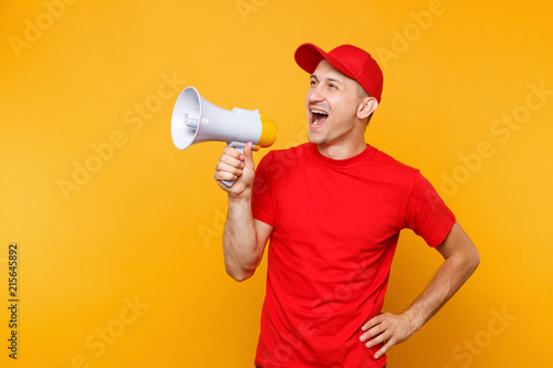 Delivery service man in red uniform isolated on yellow background. Male employee courier in cap, t-shirt screaming in megaphone hot news. Fun guy announces discounts sale. Copy space for advertisement