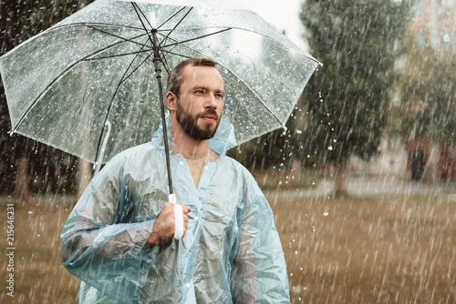 Waist up portrait of thoughtful male meditating outside. He is standing in park holding umbrella in hands looking distantly in considerations photo