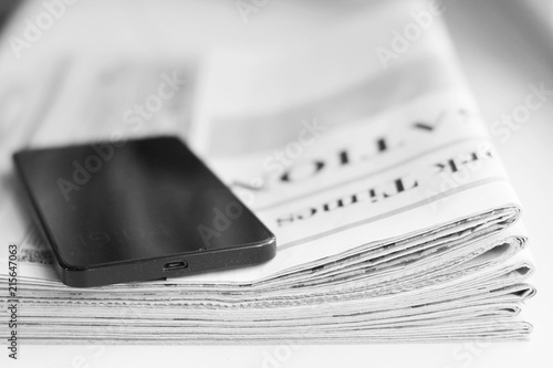 Newspaper and smartphone. Daily papers with news and black mobile phone with touch screen. Modern gadget and folded journal with articles. Electronic device on top of stacked pages, selective focus   