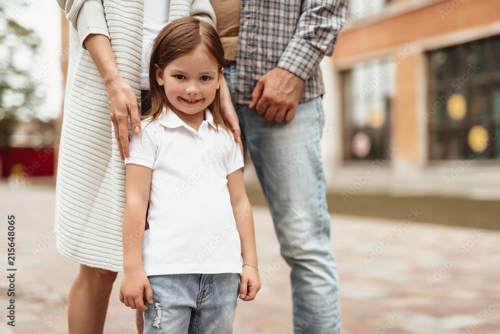 Waist up portrait of small girl standing by mother and father and smiling. They are walking on street and enjoying time outdoors together