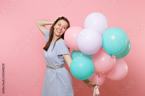 Portrait of tender smiling young woman in blue dress holding colorful air balloons keeping hand near head isolated on bright pink background. Birthday holiday party, people sincere emotions concept.