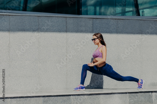 Healthy lifestyle and sport concept. Young fit female runner does stretching exercises after running in morning, shows her flexible perfect body, wears comfortable sneakers, shades and purple top