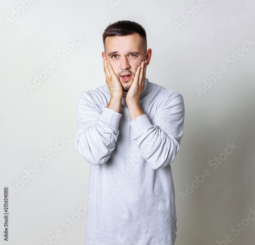 Studio portrait of adult european man holding hands near face, expressing shock and fright, dressed in a white turtleneck over gray background, copy space