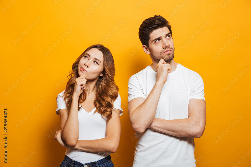 Image of happy young people man and woman in basic clothing thinking and touching chin while looking aside, isolated over yellow background - obrazy, fototapety, plakaty 