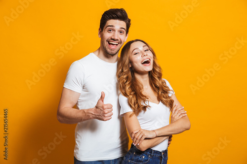Portrait of cheerful people man and woman in basic clothing smiling and showing thumb up at camera, isolated over yellow background