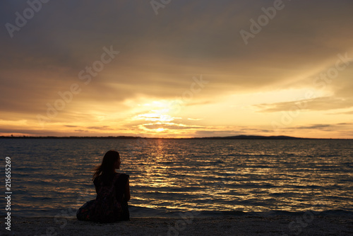 Back view silhouette of female tourist with backpack sitting alone on seashore at water edge, enjoying beautiful view of sunset on dark evening sky background. Tourism and vacations concept. © anatoliy_gleb