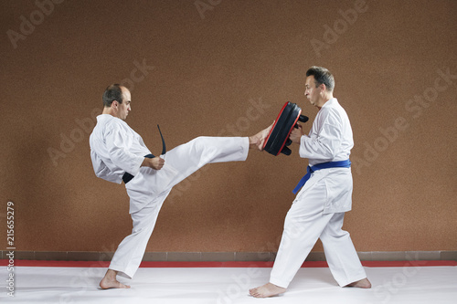 An athlete with a blue belt holds in his hands a simulator that is hit by an athlete with a black belt