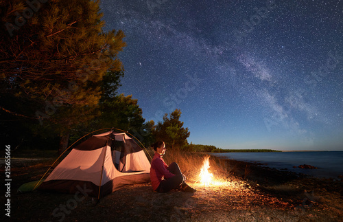 Night camping on sea shore. Smiling woman hiker sitting relaxed in front of tourist tent at campfire under amazing sky full of stars and Milky way. Clear blue water and green forest on background