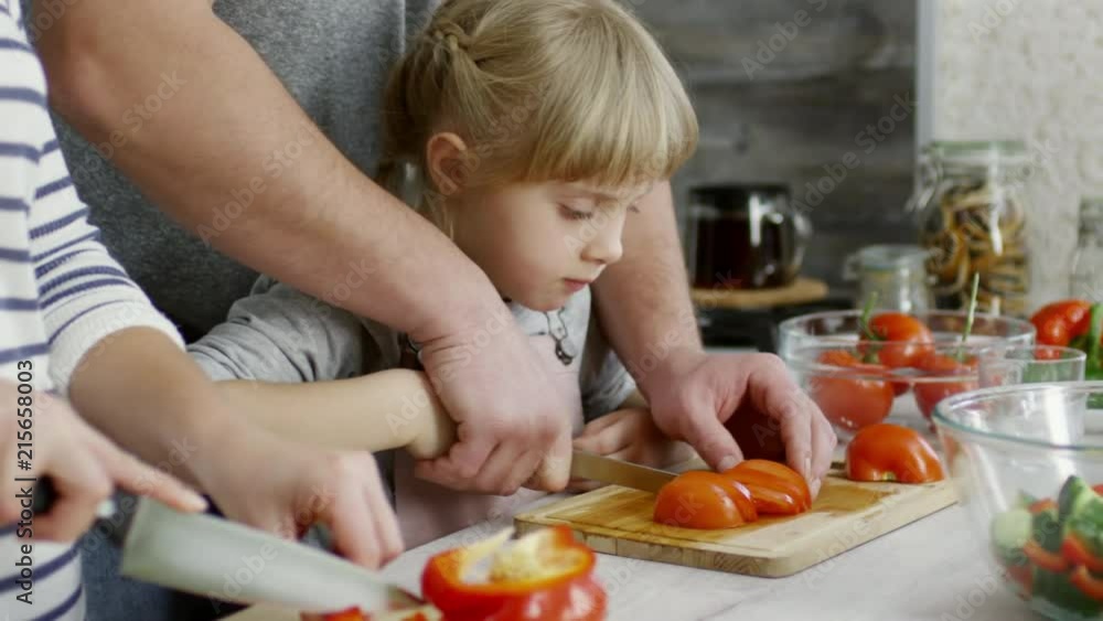 Close-up of cute little girl cutting tomato with help of father and eating one slice while cooking salad with parents Close-up of cute little girl cutting tomato with help of father and eating one slice while cooking salad with parents
