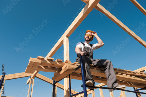 The man builder sits on the edge of the roof of the frame house, in a yellow helmet and gray overalls. The blue sky and clear sunny day.