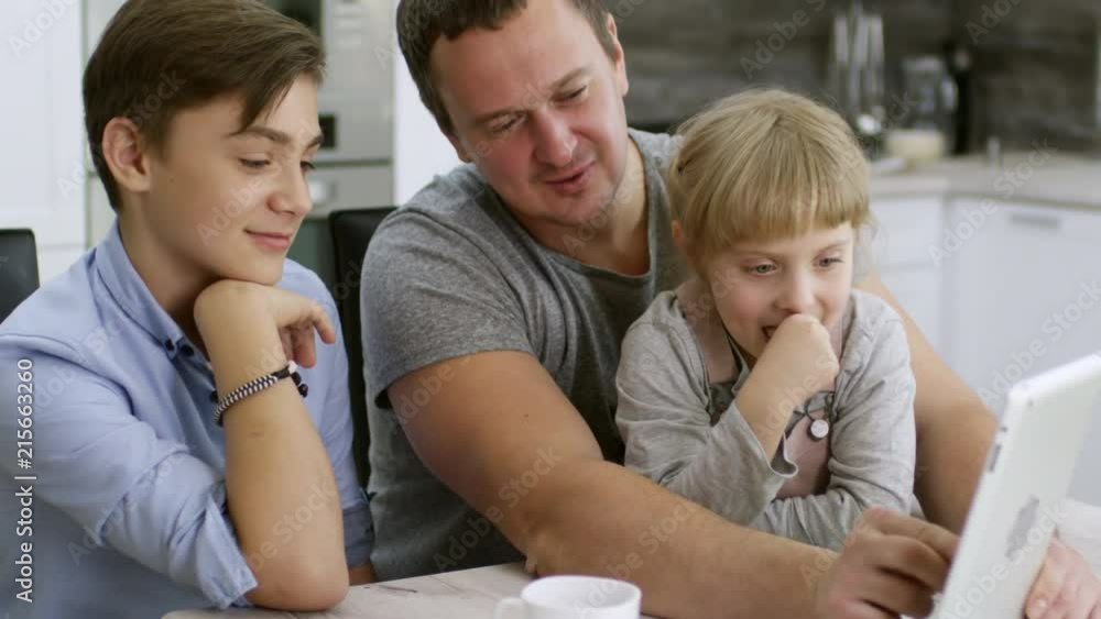 Cheerful father enjoying game on digital tablet with little daughter and teen son: family sitting at kitchen table, smiling and laughing while girl making face at front camera Cheerful father enjoying game on digital tablet with little daughter and teen son: family sitting at kitchen table, smiling and laughing while girl making face at front camera